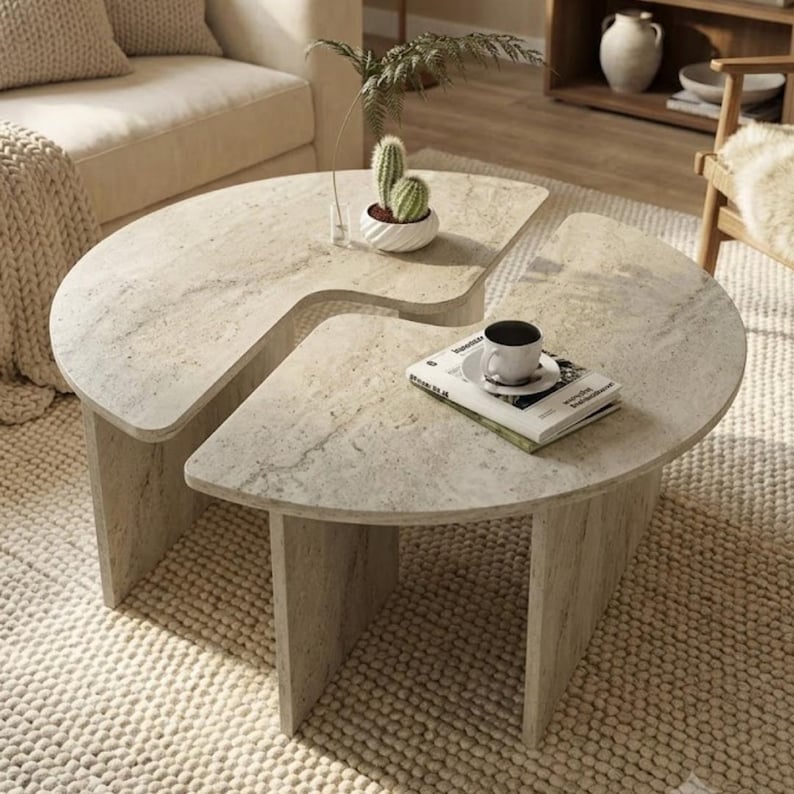 May include: A two-piece coffee table set featuring a light beige stone-like finish with natural veining. The tables are semi-circular in shape with vertical slab legs. A small potted cactus and a book with a white coffee cup sit on top.