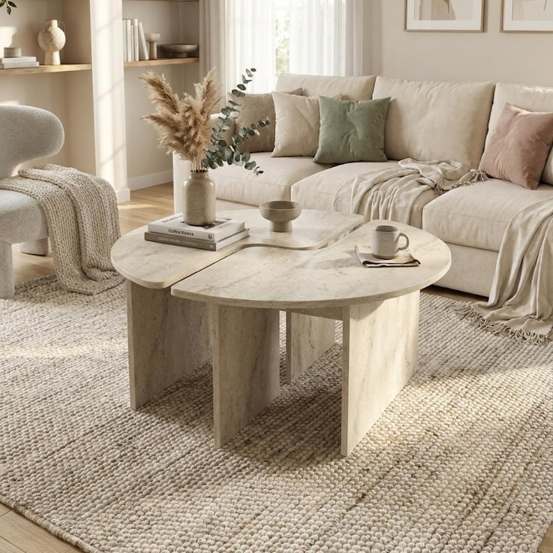 May include: A light beige, semi-circular coffee table with a smooth, stone-like finish and vertical panel legs. The table is composed of two separate pieces that fit together to form a larger surface, featuring a neutral, textured appearance.