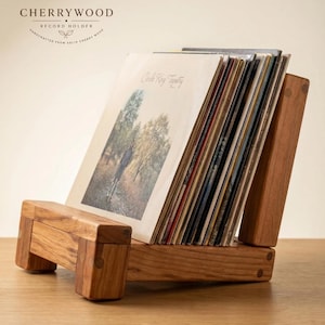 May include: A solid cherry wood record holder featuring a slanted base and back support, designed to display a collection of vinyl records. The wood has a natural grain and warm brown tone, with visible joinery details on the frame structure.