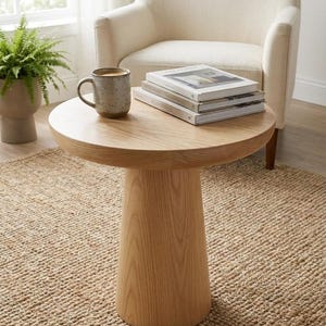 Peut inclure: Une table d'appoint ronde en bois clair avec un pied central épais. Le plateau de la table supporte une tasse en céramique mouchetée remplie d'une boisson sombre et une pile de trois livres reliés aux dos blancs avec du texte visible.