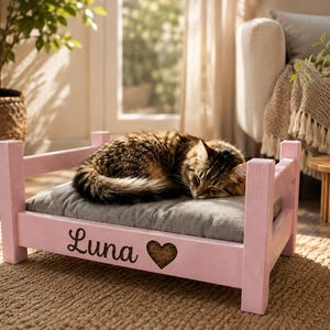 May include: A small, light pink wooden pet bed frame with four corner posts and a gray cushioned mattress. The front panel features the name Luna in black cursive script followed by a dark brown heart-shaped cutout. A tabby cat is sleeping.