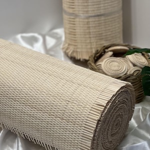 May include: A rolled-up, natural-colored woven mat made of thin, parallel reeds. In the background, a tall, cylindrical woven basket and a small basket filled with decorative wooden pieces are visible. The items are on a white, satin-like surface.