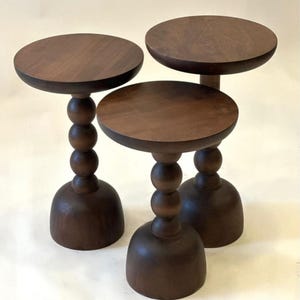 Puede incluir: Tres mesas auxiliares de madera marrón oscuro. Cada mesa tiene una tapa redonda, un tallo con tres esferas y una base en forma de campana. Las mesas están dispuestas en grupo, mostrando su diseño y artesanía.