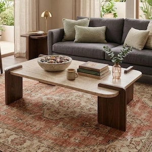 May include: A rectangular coffee table with a light beige stone top and dark brown wooden legs. On the table sits a large stone bowl filled with smooth pebbles, a small ceramic mug, and a stack of three hardcover books next to a glass vase.