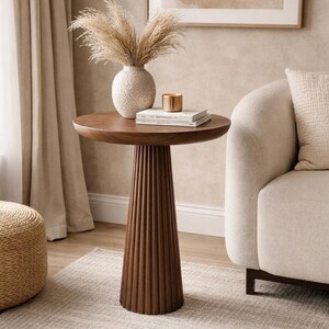 May include: A round wooden side table with a dark brown finish and a fluted, pedestal-style base. The tabletop is smooth and circular, supported by a thick, vertically ribbed column that tapers slightly towards the floor, creating a stable structure.