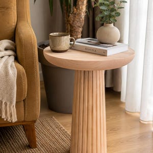 Peut inclure: Une table d'appoint en bois clair avec un plateau circulaire lisse et une base cylindrique cannelée. La table supporte une tasse en céramique mouchetée, un livre intitulé Kinfolk et un petit vase contenant des tiges d'eucalyptus.