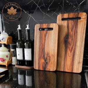 Puede incluir: Dos tablas de cortar rectangulares de madera con vetas naturales y ranuras para asas integradas, junto a dos botellas de aceite de vidrio oscuro con etiquetas blancas y vertedores plateados, y un tarro de cer&aacute;mica blanco con flores.