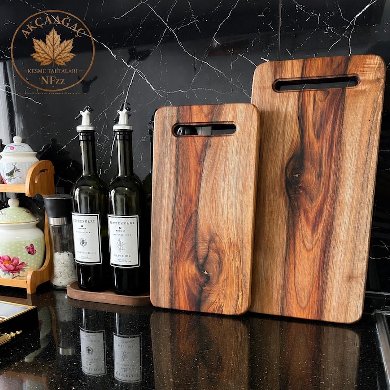 Puede incluir: Dos tablas de cortar rectangulares de madera con vetas naturales y ranuras para asas integradas, junto a dos botellas de aceite de vidrio oscuro con etiquetas blancas y vertedores plateados, y un tarro de cer&aacute;mica blanco con flores.