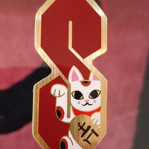 May include: A red and gold sticker featuring a stylized letter "S" with a lucky cat illustration. The cat is white with black markings, a red collar, and holds a gold coin with Japanese characters.