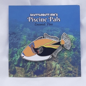 May include: An enamel pin featuring a colorful fish design. The pin has a white, orange, black, and yellow color scheme. The pin is on a blue card with the text "SCUTTLEBUTT INK'S Piscine Pals Enamel Pins". The background of the card shows an underwater scene.