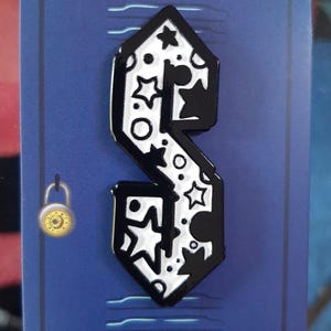 May include: A black and white enamel pin in the shape of the stylized "S" design, featuring stars and circles. The pin is on a blue background with a gold-colored padlock illustration.