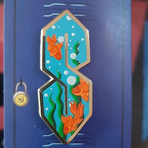 May include: Enamel pin featuring a stylized 'S' design with a turquoise interior, gold trim, and three orange goldfish swimming in a blue water background. The pin is set against a blue background with a gold padlock.