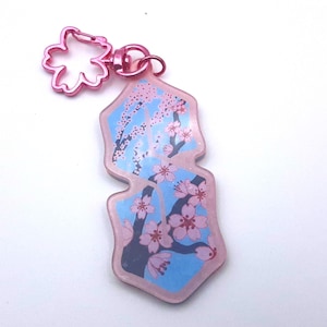 Op de afbeelding: Een roze sleutelhanger met een kersenbloesem ontwerp. De sleutelhanger heeft een lichtblauwe achtergrond met roze kersenbloesemtakken en bloemen. Het heeft een roze bloemvormige sluiting.