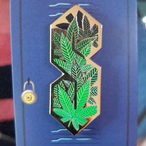 May include: A gold-coloured pin in the shape of the letter "S" with a cannabis leaf design. The pin features green and black leaves, set against a blue background with a lock and key illustration.
