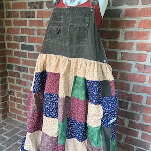 May include: A long, olive green corduroy overall dress with a patchwork skirt. The skirt features squares of floral, solid, and patterned fabrics in shades of blue, green, burgundy, and cream. The dress is displayed on a mannequin.