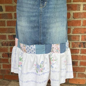 May include: A denim skirt with a patchwork ruffle. The skirt features a blue denim upper section and a lower section with a patchwork design of floral and patterned fabrics in shades of blue, pink, and white. The skirt is displayed on a white stand.