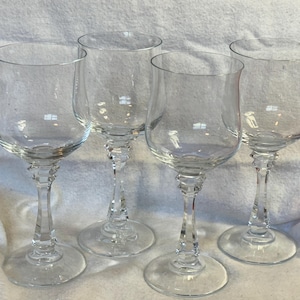 May include: Four clear glass wine goblets with a tulip-shaped bowl and a decorative stem. The goblets are made of clear glass and have a round base. The stem has a decorative design. The goblets are arranged on a white surface.