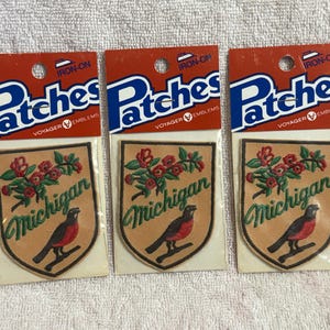 May include: Three iron-on patches featuring a shield design with the word "Michigan" in green script, a red robin, and red roses. The patches are on a tan background and are packaged on a red and blue card that reads "Patches".