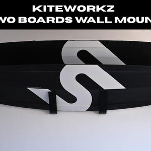 Può includere: Tavole da kitesurf in bianco e nero montate a parete. Le tavole presentano un grande design a "S" bianco e dettagli turchese. Il testo "KITEWORKZ TWO BOARDS WALL MOUNT" è visualizzato sopra le tavole.