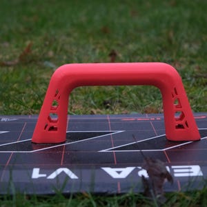 Könnte beinhalten: Ein leuchtend roter Kunststoffgriff mit einem Ausschnittmuster, montiert auf einer schwarz-weißen Oberfläche. Der Griff ist U-förmig und hat eine glatte, abgerundete Oberseite.