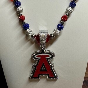 Puede incluir: Un collar de cuentas rojas, blancas y azules con un dije de plata con la letra "A" con un halo sobre ella. El dije est&aacute; cubierto de diamantes de imitaci&oacute;n.