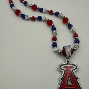 Puede incluir: Un collar de cuentas rojas, blancas y azules con un colgante plateado que presenta el logotipo del equipo de b&eacute;isbol de los &Aacute;ngeles de Los &Aacute;ngeles. El colgante est&aacute; cubierto de diamantes de imitaci&oacute;n.