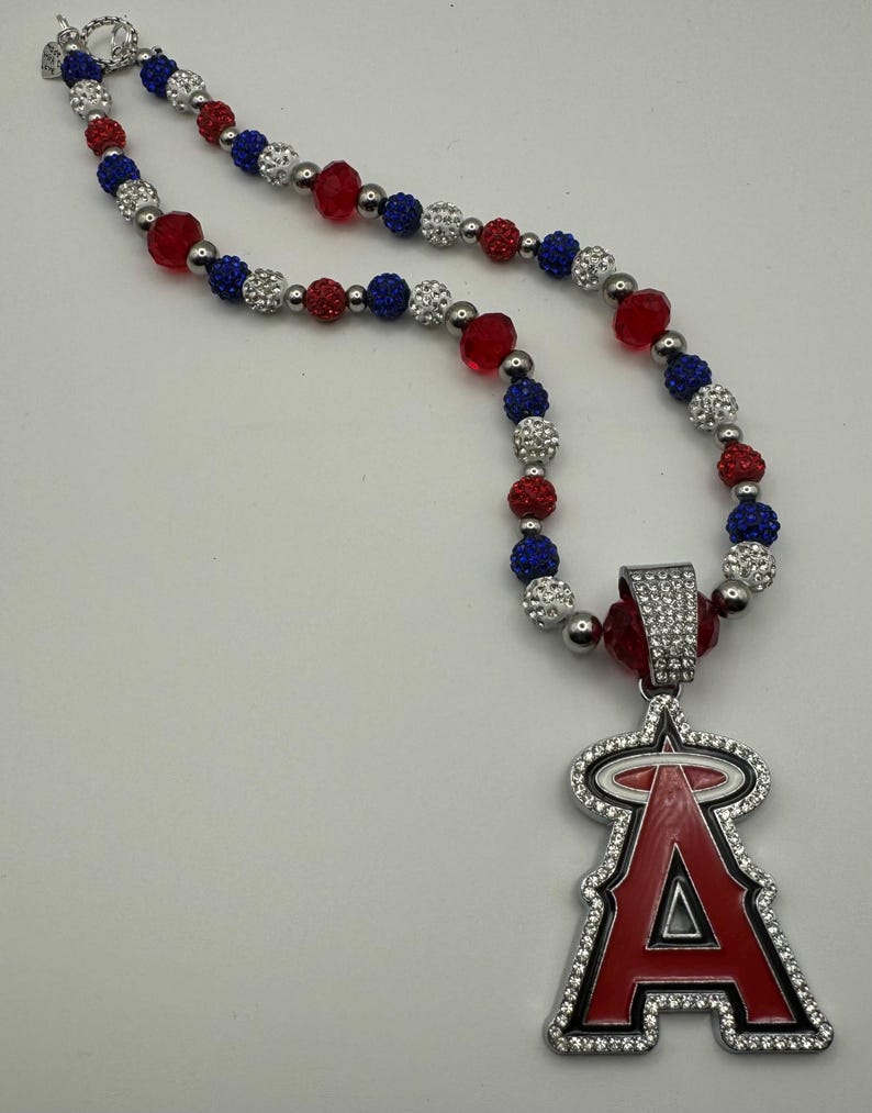 Puede incluir: Un collar de cuentas rojas, blancas y azules con un colgante plateado que presenta el logotipo del equipo de b&eacute;isbol de los &Aacute;ngeles de Los &Aacute;ngeles. El colgante est&aacute; cubierto de diamantes de imitaci&oacute;n.