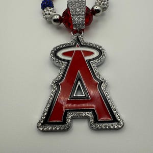 Puede incluir: Un collar de cuentas rojas, blancas y azules con un colgante plateado que presenta el logotipo del equipo de b&eacute;isbol de los &Aacute;ngeles de Los &Aacute;ngeles. El colgante est&aacute; adornado con diamantes de imitaci&oacute;n.
