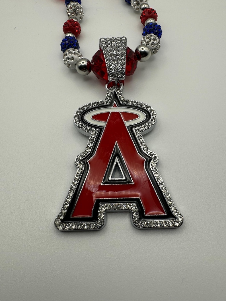 Puede incluir: Un collar de cuentas rojas, blancas y azules con un colgante plateado que presenta el logotipo del equipo de b&eacute;isbol de los &Aacute;ngeles de Los &Aacute;ngeles. El colgante est&aacute; adornado con diamantes de imitaci&oacute;n.