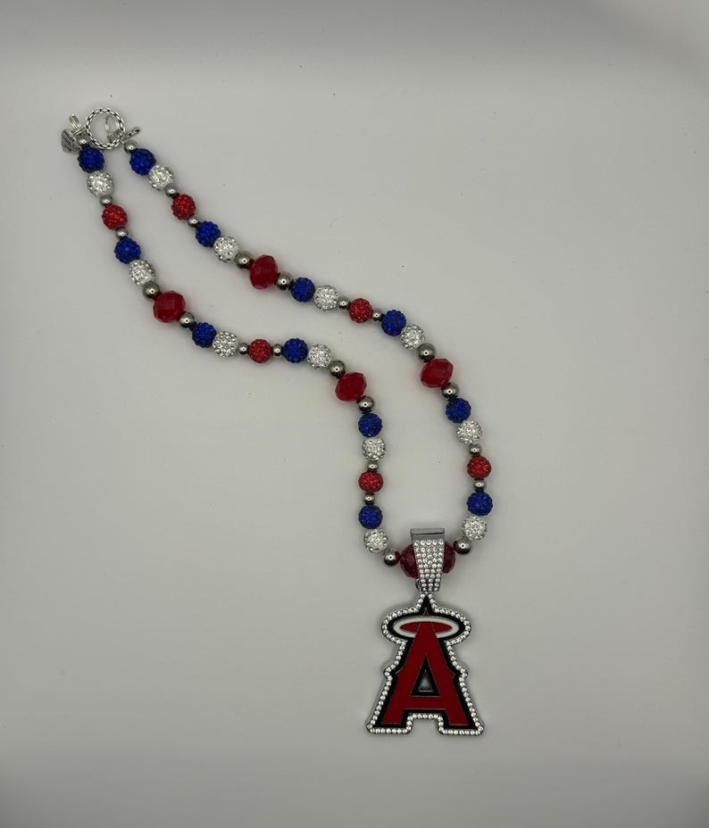 Puede incluir: Un collar de cuentas rojas, blancas y azules con un colgante plateado que presenta el logotipo del equipo de b&eacute;isbol de los &Aacute;ngeles de Los &Aacute;ngeles.