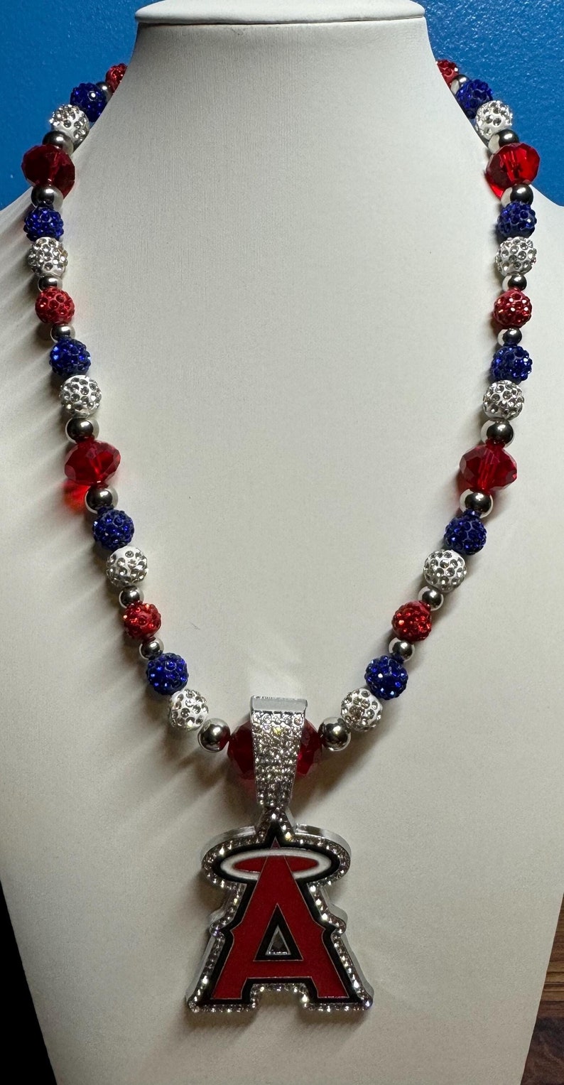 Puede incluir: Un collar de cuentas rojas, blancas y azules con un dije de plata con la letra "A" con un halo sobre ella. El dije est&aacute; cubierto de diamantes de imitaci&oacute;n.