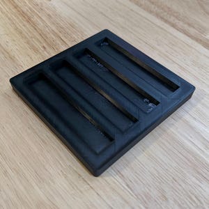 May include: A black, square organizer with three rectangular slots. The organizer is made of a textured material and sits on a wooden surface. The slots are designed to hold small items.