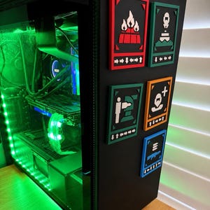 May include: A custom-built computer case with green LED lighting. Six colorful, rectangular game-themed icons are mounted on the side. The icons feature white symbols on red, green, orange, and blue backgrounds. The computer case has a clear side panel.