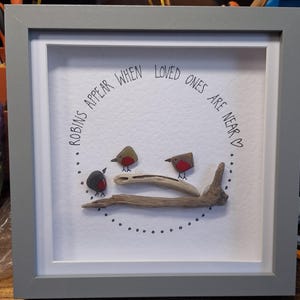 May include: A framed artwork with a white background featuring a quote about robins appearing when loved ones are near. The quote is surrounded by a circle of small black dots. Three small painted robins are perched on a piece of driftwood.
