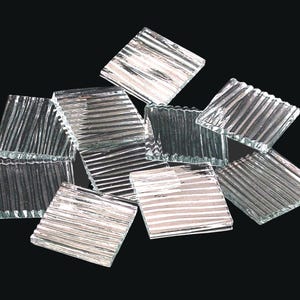 May include: A collection of clear, square glass tiles with a textured, ridged surface. The tiles are arranged in a scattered pattern against a black background. The glass has a reflective quality, catching the light and creating highlights.