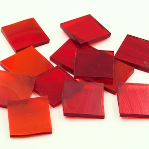 May include: A collection of square, translucent glass tiles in shades of red, from light orange-red to deep crimson. The tiles are scattered on a white surface, showcasing their varying hues and textures. Ideal for mosaic art.