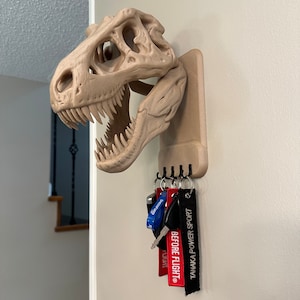 May include: A beige 3D printed dinosaur skull key holder mounted on a wall with a set of keys and keychains hanging from it. The keychains are black with white text that reads "Before Flight" and "Tanaka Power Sport".