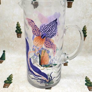 Puede incluir: Una jarra de vidrio transparente con asa, decorada con ilustraciones florales e insectos coloridos. El dise&ntilde;o presenta flores naranjas, moradas y rosas, junto con una mariposa o polilla. La jarra es adecuada para servir bebidas.