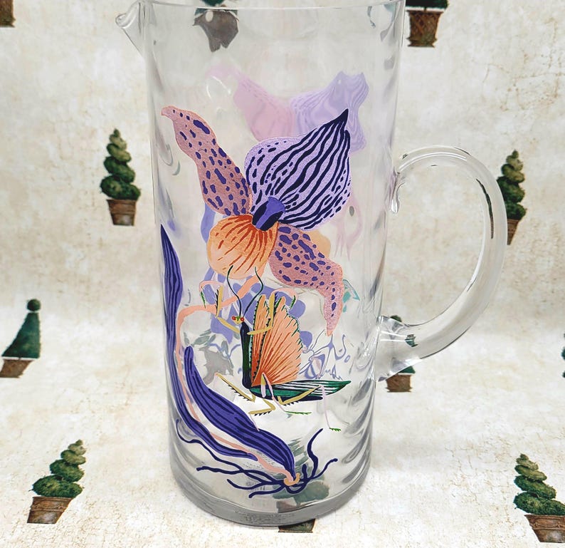Puede incluir: Una jarra de vidrio transparente con asa, decorada con ilustraciones florales e insectos coloridos. El dise&ntilde;o presenta flores naranjas, moradas y rosas, junto con una mariposa o polilla. La jarra es adecuada para servir bebidas.