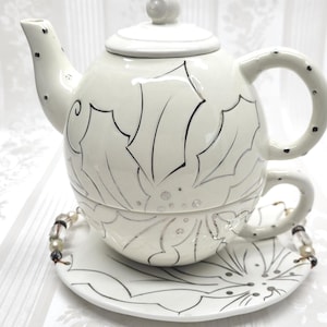 May include: A white ceramic tea set with a teapot, teacup, and saucer. The set features a black leaf design and black polka dots. The saucer has a beaded handle. The teapot has a lid and spout.