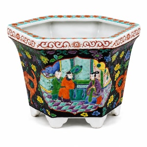 May include: A hexagonal ceramic planter featuring a black background with colorful floral patterns and a central cartouche depicting three figures in traditional attire. The rim has a red and white scroll design, and the base is white with six feet.