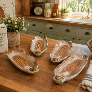 May include: Four ceramic corn-shaped serving dishes with a tan and cream color scheme are displayed on a wooden table. The dishes have a detailed design resembling corn on the cob. A rustic kitchen setting includes a jar with "Farmer's Market" text.