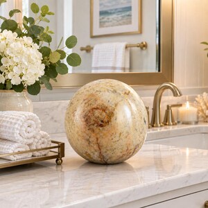 May include: A smooth, spherical decorative object made of polished stone with a variegated pattern of cream, beige, and brown tones. The sphere features natural veining and mineral inclusions throughout its surface, creating a unique, organic appearance.