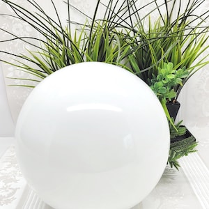 May include: A large, glossy white sphere, possibly a decorative light fixture or globe, sits on a small base. The sphere is the focal point, reflecting light. Green plants in black pots are in the background, adding a natural contrast.