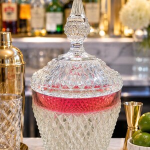 May include: A clear glass punch bowl with a matching lid, featuring a diamond-cut pattern across the exterior. The bowl has a wide, horizontal band of translucent red glass near the top rim, and the lid is topped with a pointed, faceted finial.