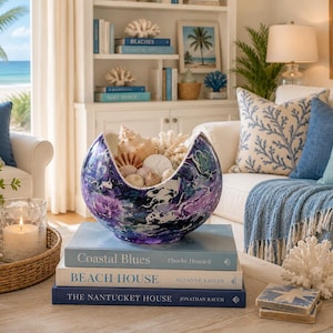 May include: A round, open-top decorative bowl with a marbled purple, blue, and white finish, filled with various seashells and coral pieces, resting on a stack of three hardcover books titled Coastal Blues, Beach House, and The Nantucket House.