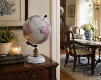 May include: A decorative globe featuring a muted color palette of grey, white, and dusty rose continents. The globe is mounted on a dark wooden stand with a circular white base and includes a small black finial at the top of the axis.