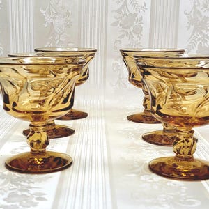 May include: Six amber-colored, stemmed dessert glasses. Each glass features a decorative, swirling pattern on the bowl and stem. The glasses are arranged in two rows of three, showcasing their elegant design.