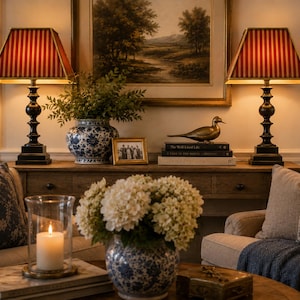 May include: Two table lamps with black bases and red and gold striped rectangular shades, a blue and white floral ceramic vase, a brass bird figurine, a framed photograph, and a glass candle holder with a white pillar candle on a wooden table.