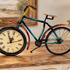 May include: A decorative metal bicycle sculpture featuring a functional analog clock face in the front wheel. The frame is painted in distressed shades of blue, yellow, and red. The clock face has a cream background with black Roman numerals and the word QUARTZ printed in black.
